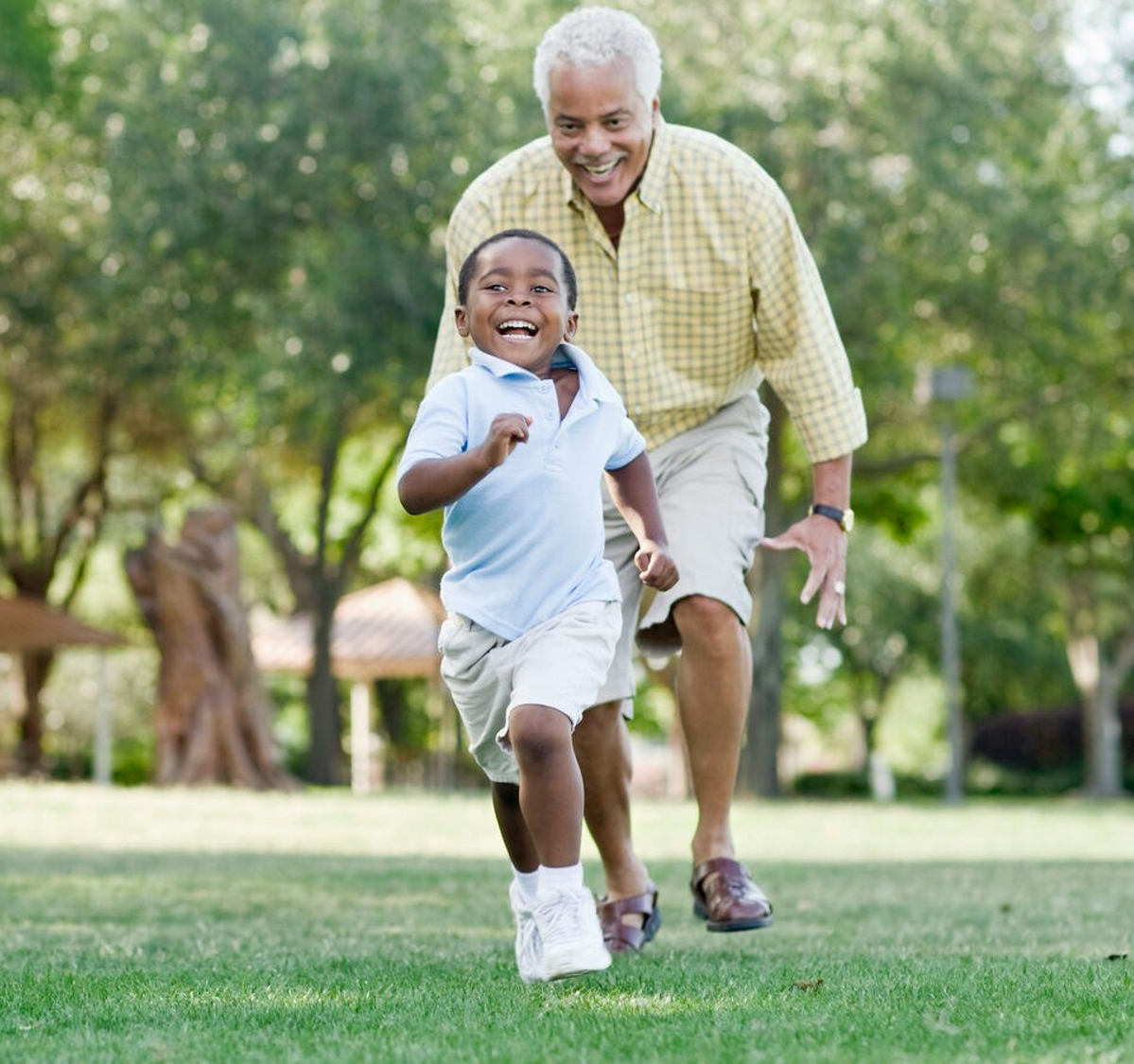 A grandfather joyfully chasing his laughing grandson across a sunny park lawn